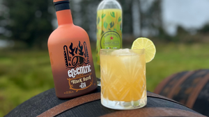 A Dark and Stormy cocktail sitting on a barrel outside next to a bottle of Black Batch Spiced Rum and a bottle of Limecello.