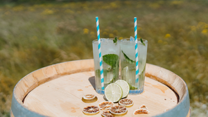 Two high-ball glasses of our Ginger Spiced Mojito cocktail sitting on a barrel outside in the sunshine, with a field of grass and wildflowers in the background.
