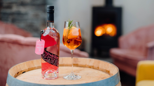 A bottle of our refreshing Pembrokeshire Pinky Welsh Gin next to a Pinky Aperol Spritz cocktail in a delicate wine glass on a barrel with a fire in the background.
