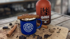 A Spiced Rum Hot Chocolate sitting on a wooden board surrounded by cinnamon sticks, chocolate pieces and whole cloves, with a bottle of Black Batch Spiced Rum in the background.
