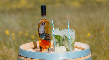 A bottle of our Black Batch Welsh Rum, a whole lime cut in half, and two high-ball glasses of our Ginger Spiced Mojito cocktail sitting on a barrel outside in the sunshine, with a field of grass and wildflowers in the background.