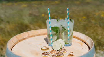 Two high-ball glasses of our Ginger Spiced Mojito cocktail sitting on a barrel outside in the sunshine, with a field of grass and wildflowers in the background.