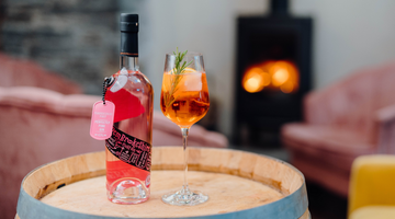 A bottle of our refreshing Pembrokeshire Pinky Welsh Gin next to a Pinky Aperol Spritz cocktail in a delicate wine glass on a barrel with a fire in the background.