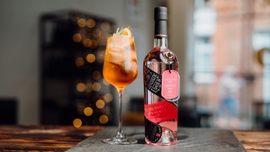 A bottle of our refreshing Pembrokeshire Pinky Welsh Gin next to a Pinky Aperol Spritz cocktail in a delicate wine glass on top of slate with fairy lights in the background.