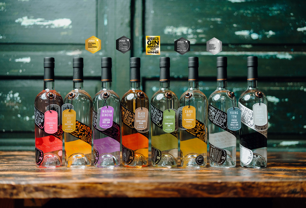 Each distinctive bottle of Welsh gin, Welsh rum and Welsh vodka in the Eccentric Spirit Co range standing side by side on a wooden table with a green, rustic background, with a variety of awards won by different Welsh spirits within the range sitting above them.